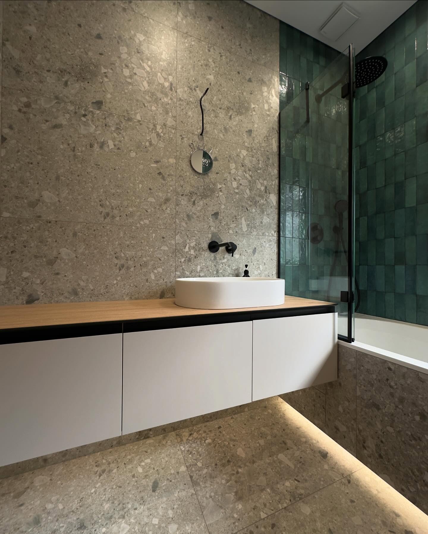 Фрагмент ванної кімнати: підвісна тумба під раковину з білими фасадами, дерев'яною стільницею та накладною білою раковиною, сіра стіна під терраццо, акцентна зелена плитка у душовій зоні.
