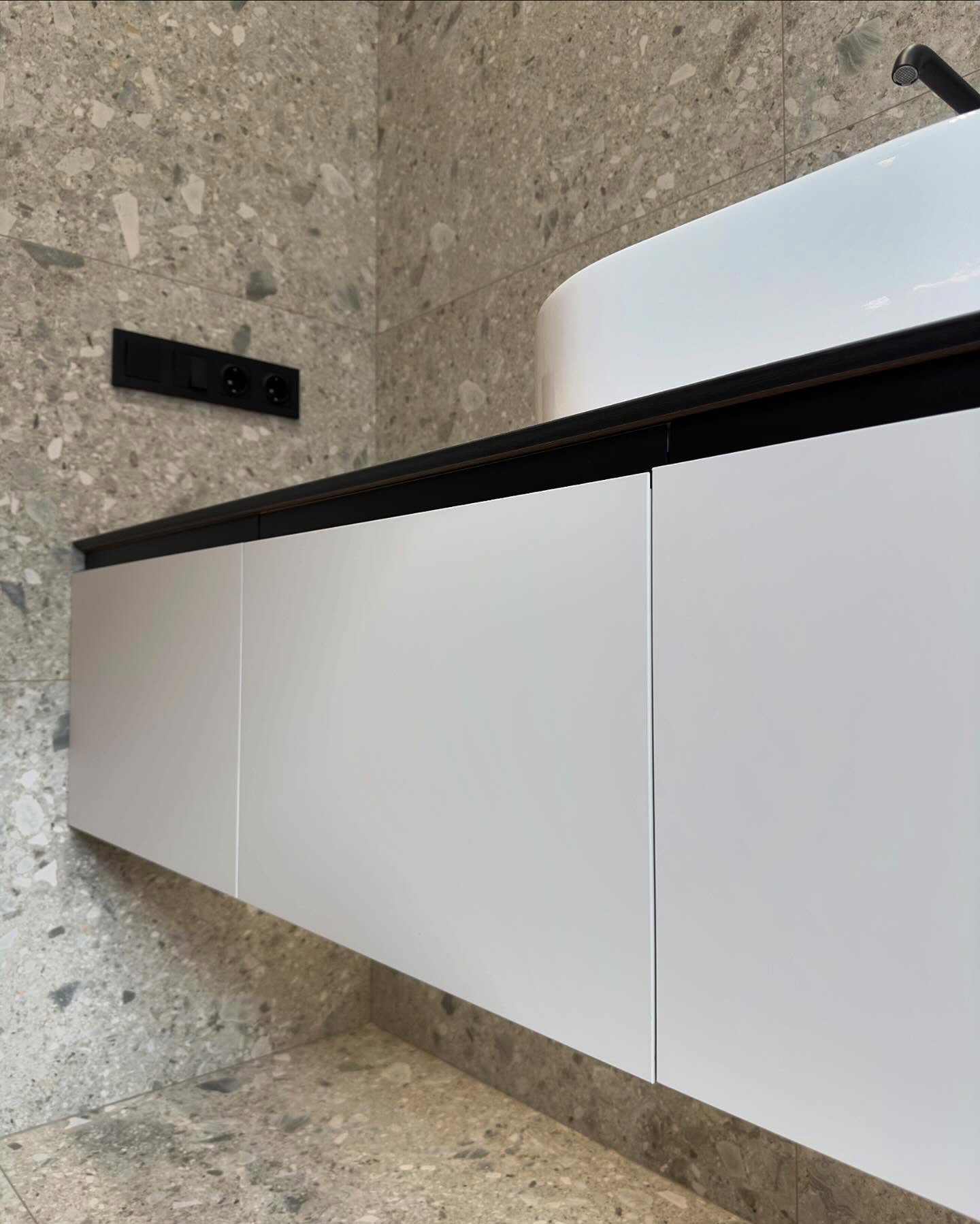 Деталь інтер'єру: накладна біла раковина на дерев'яній стільниці підвісної тумби з чорною окантовкою, чорний настінний змішувач, скляна перегородка душової зони з акцентною зеленою плиткою.