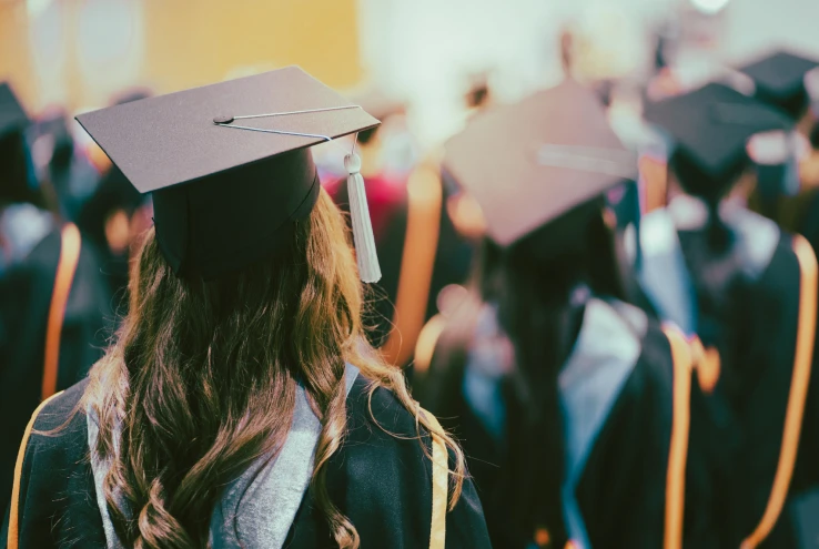 Back view of a graduate wearing a black cap and gown with long wavy hair at a commencement ceremony.