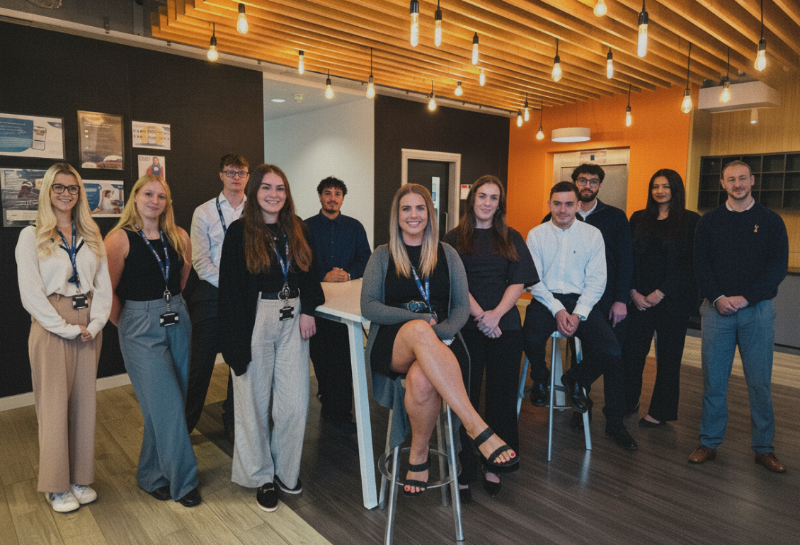 Back view of a graduate wearing a black cap and gown with long wavy hair at a commencement ceremony.