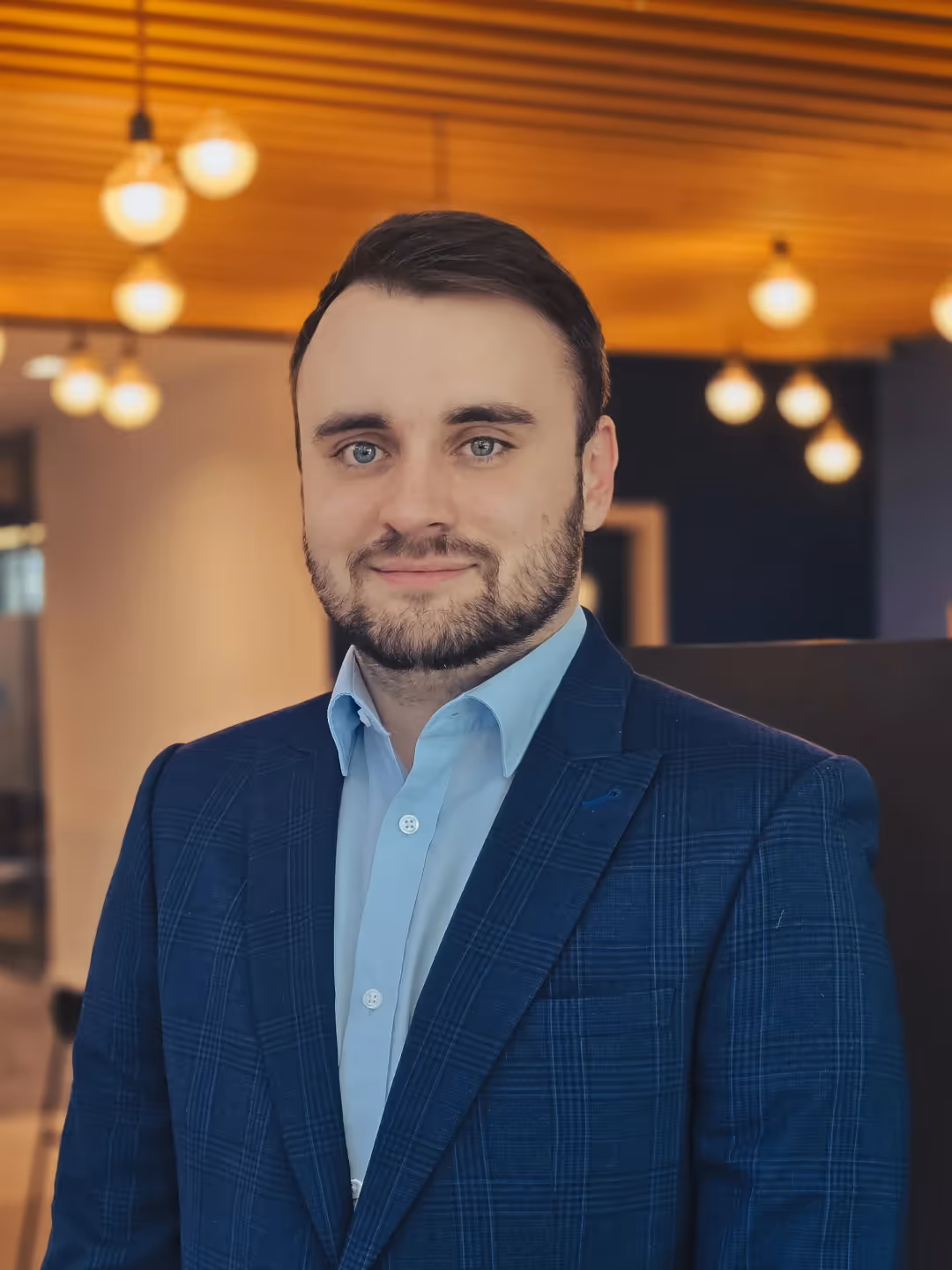 Man with short dark hair and beard in a blue checkered suit and light blue shirt indoors.