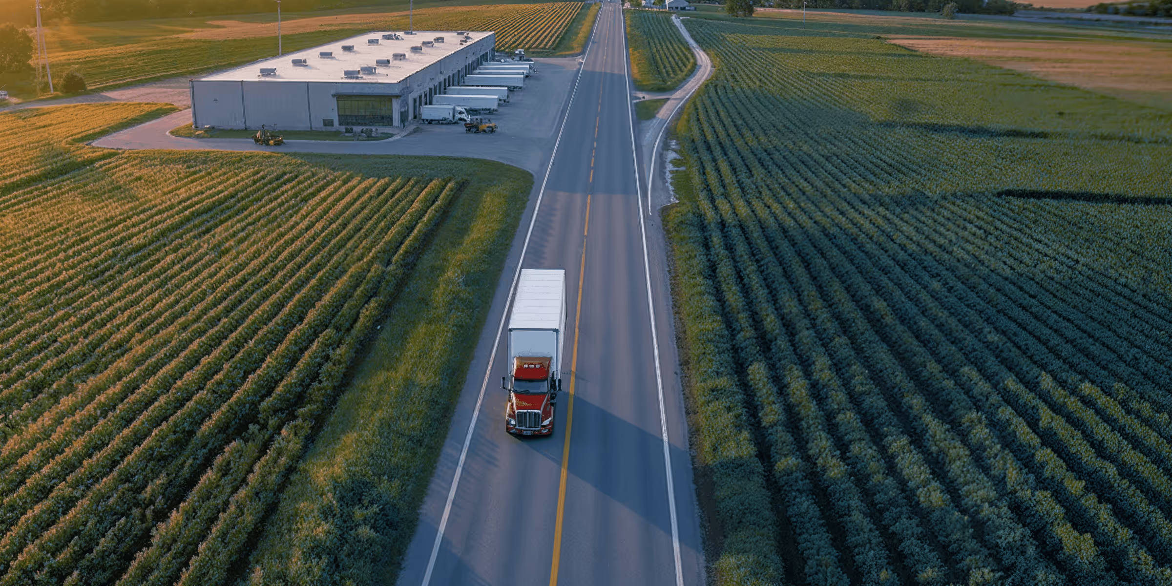 Aerial view of a red semi-truck driving on a two-lane road flanked by green farmland and a warehouse with parked trailers.