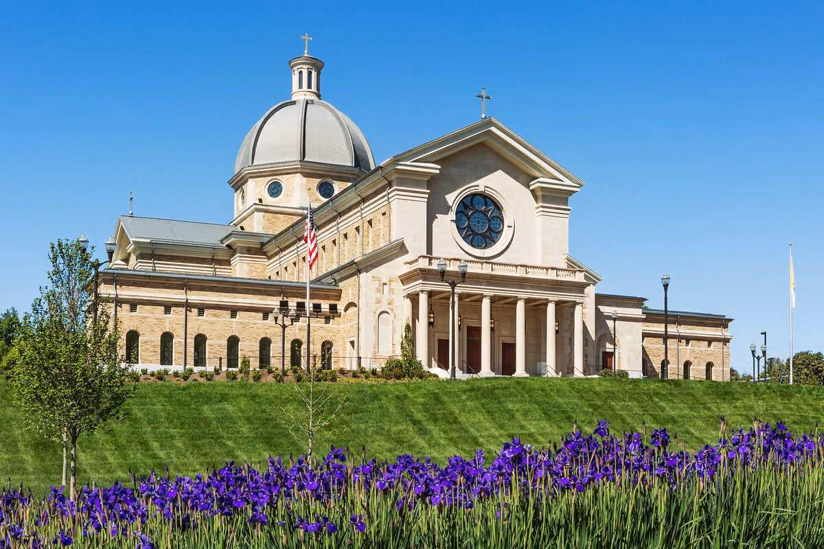 Classical church building with a large dome, columns at entrance, American flag, and purple flowers in the foreground under a clear blue sky.