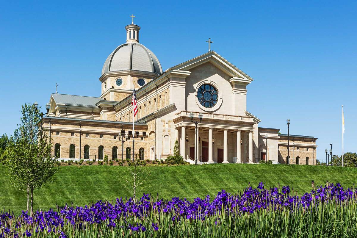 Classical church building with a large dome, columns at entrance, American flag, and purple flowers in the foreground under a clear blue sky.