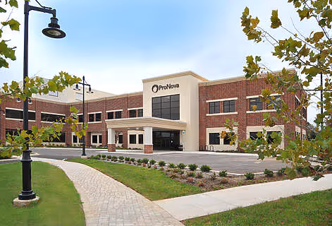 Two-story brick office building with a sign reading ProNova, surrounded by green lawn and trees under a clear sky.