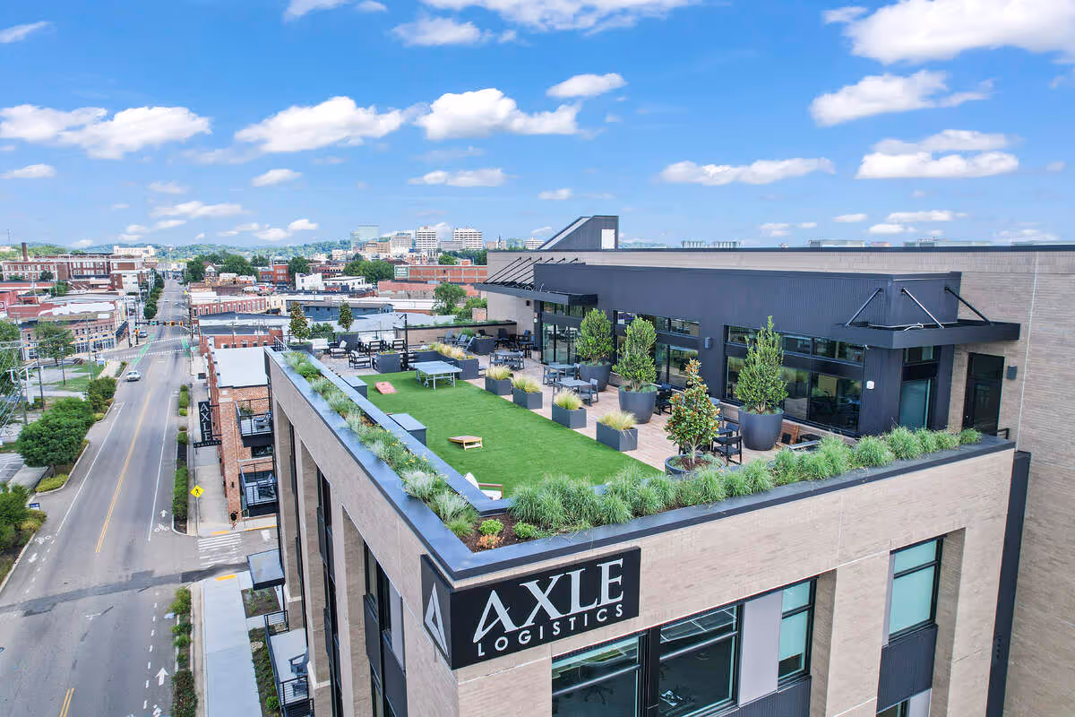 Rooftop terrace with green lawn, seating, and plants on a building labeled Axle Logistics in an urban area under a blue sky with clouds.