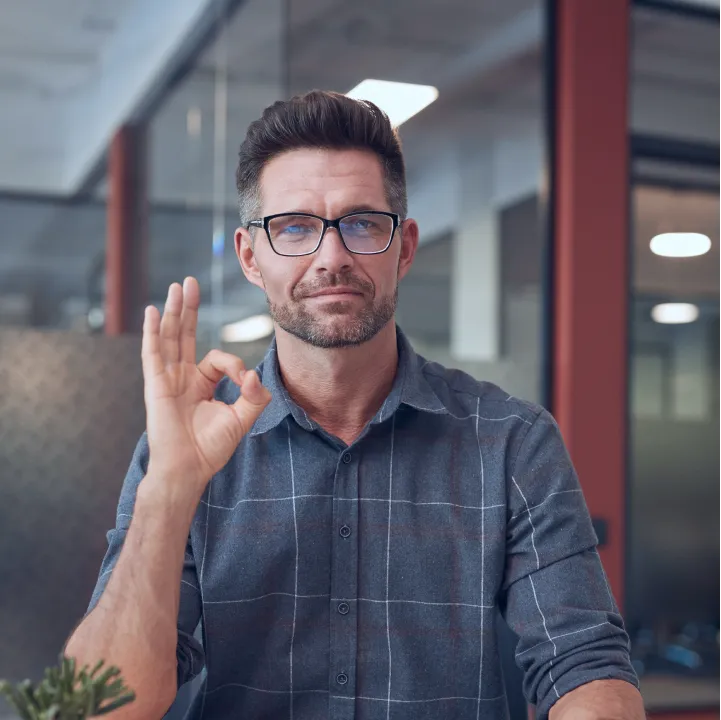 Man in glasses and checkered shirt showing an OK hand gesture in an office setting.