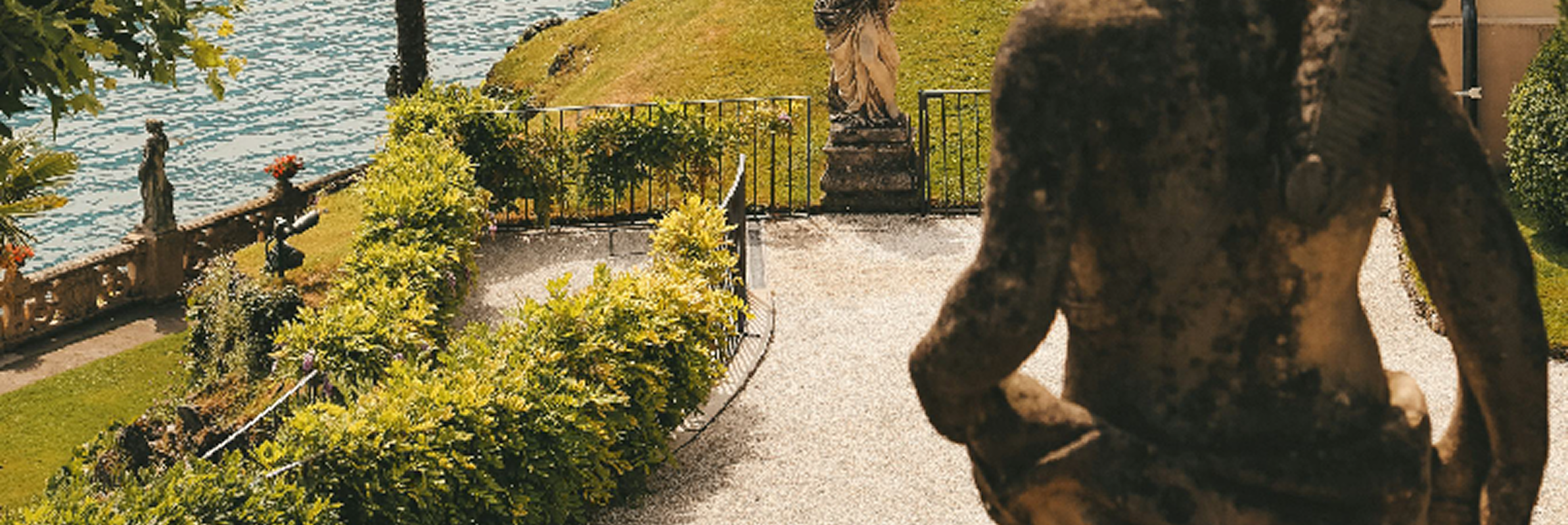Bronze or dark statue in foreground with lakeside pathway, landscaped gardens, and water view in background