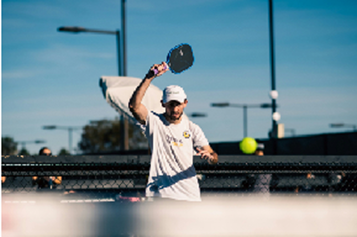 A pickleball player in white clothing and cap mid-swing with paddle raised overhead, hitting a yellow ball on an outdoor court with chain-link fencing and light poles in the background