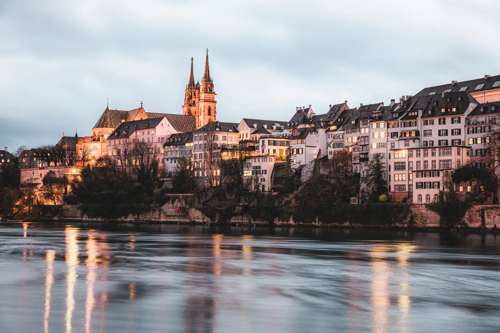Abendliche Ansicht der Basler Altstadt mit beleuchteten Häusern und dem Münster am Rhein, dessen Spiegelungen im Wasser sichtbar sind.