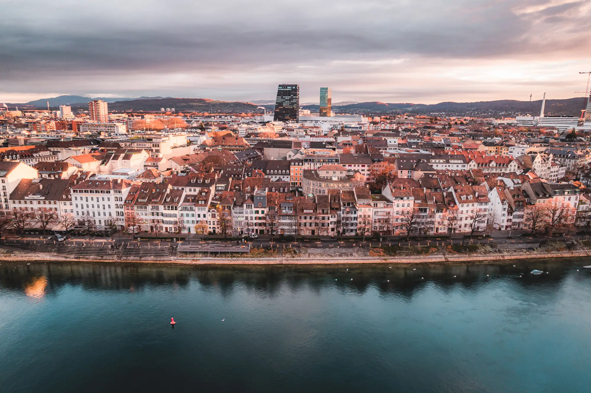 Panoramablick auf Basel mit dicht bebauter Altstadt entlang des Rheins, moderner Skyline im Hintergrund und ruhigem Fluss im Vordergrund.