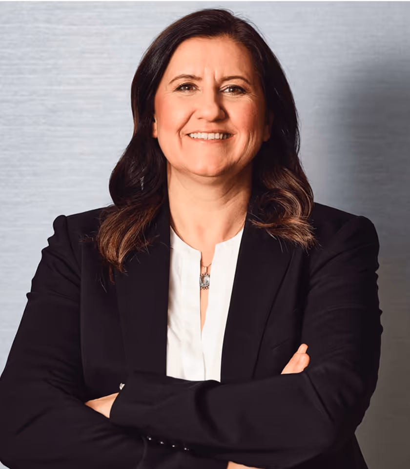 Frau mit dunklem Haar im schwarzen Blazer lächelt mit verschränkten Armen vor neutralem Hintergrund.