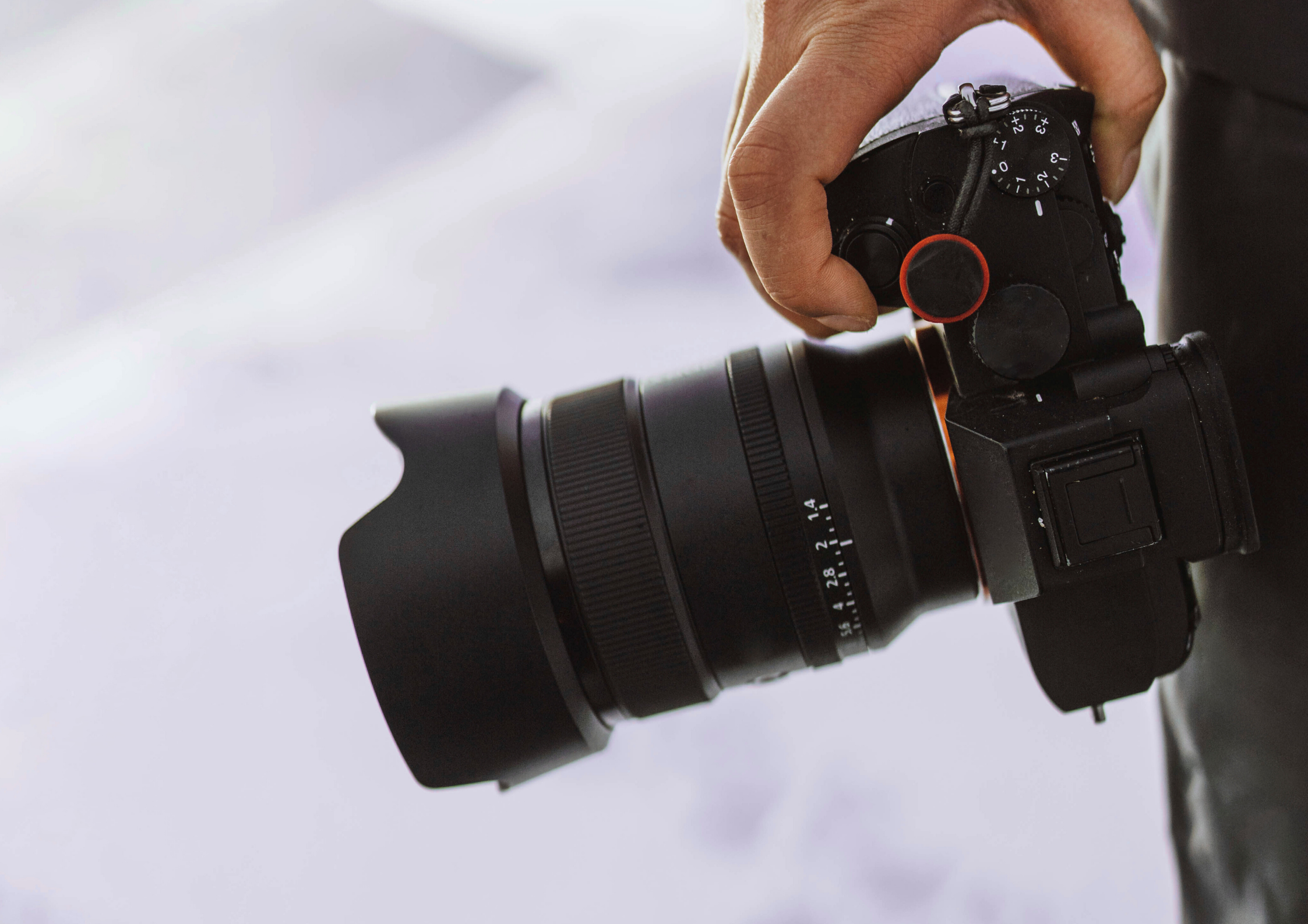 Close-up of a hand holding a black camera with a large lens and lens hood against a blurred background.