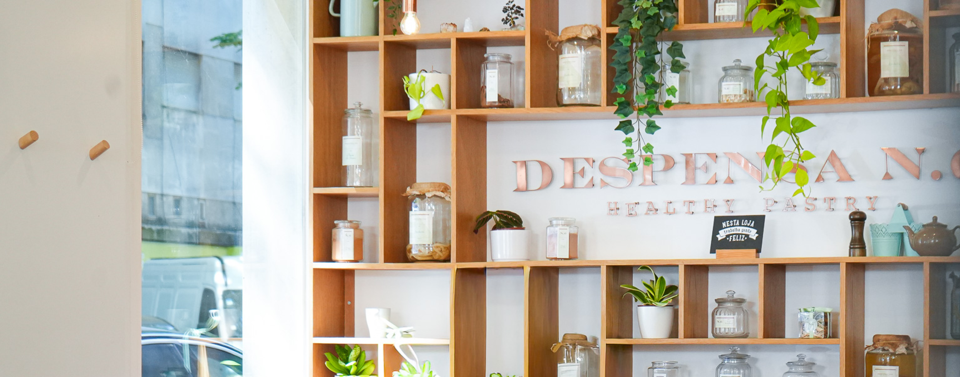 Estante de madeira com potes de vidro, plantas e decoração, com letras na parede a dizer 'DESPENSA N.C. HEALTHY PASTRY'.