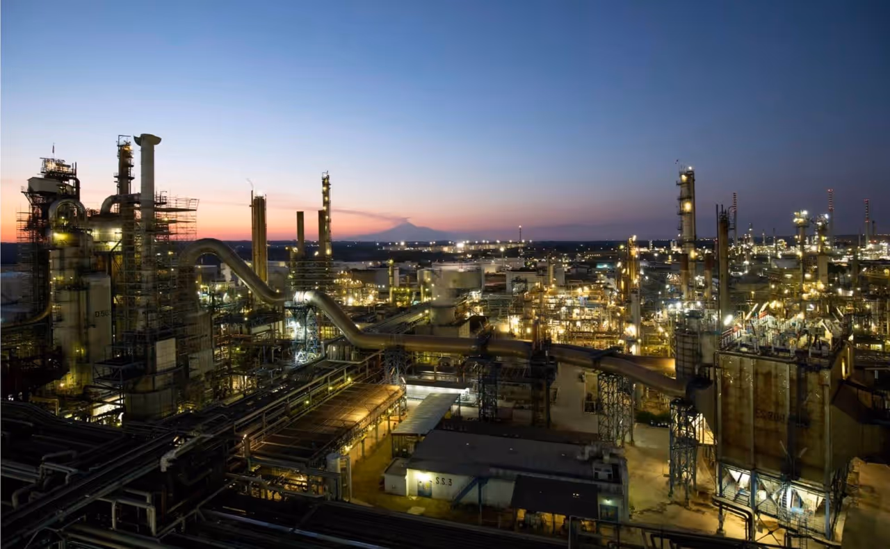 Industrial refinery complex illuminated at dusk with a clear sky and distant mountains.