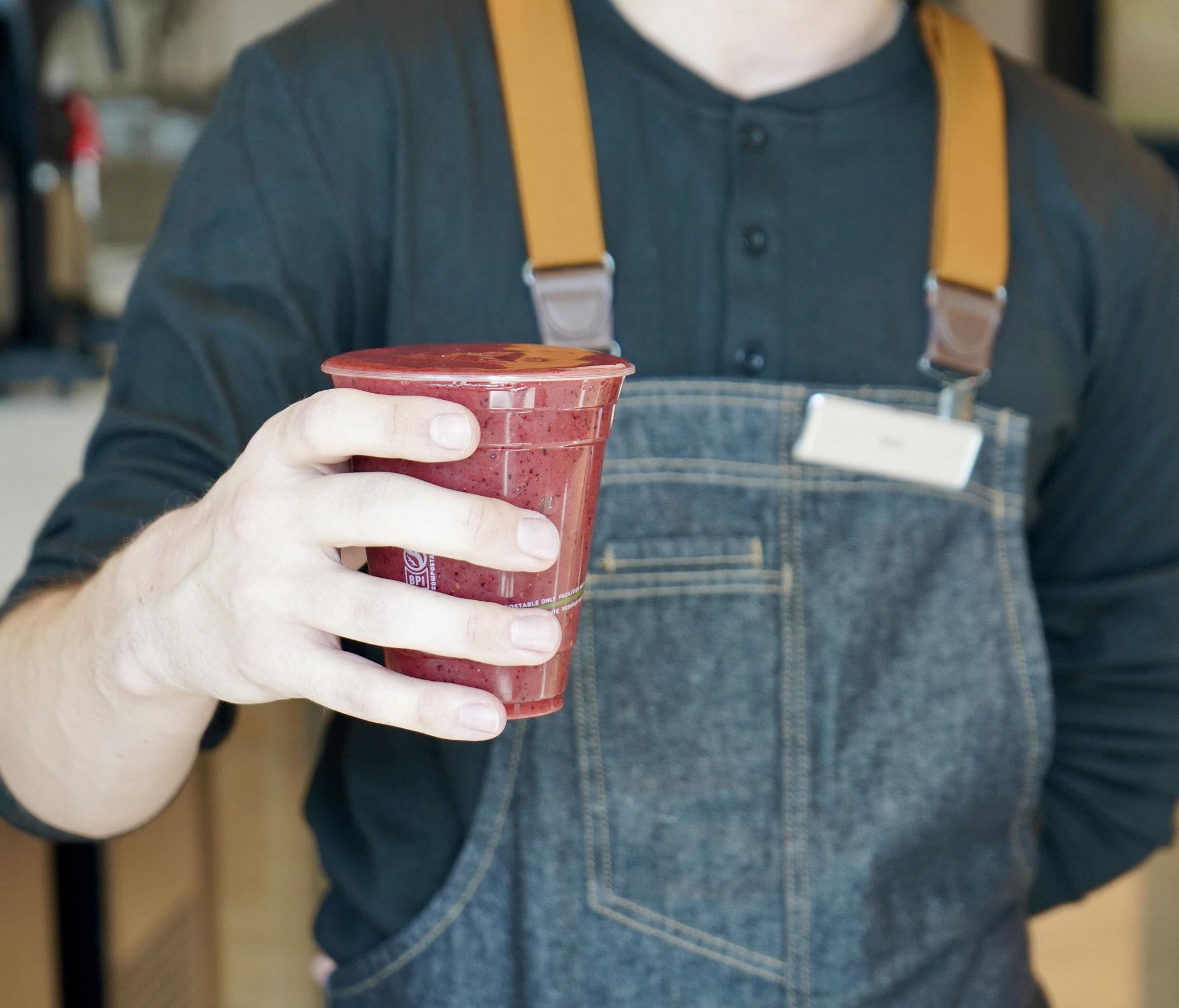 Person wearing denim apron and black shirt holding a red smoothie in a clear plastic cup with a lid.