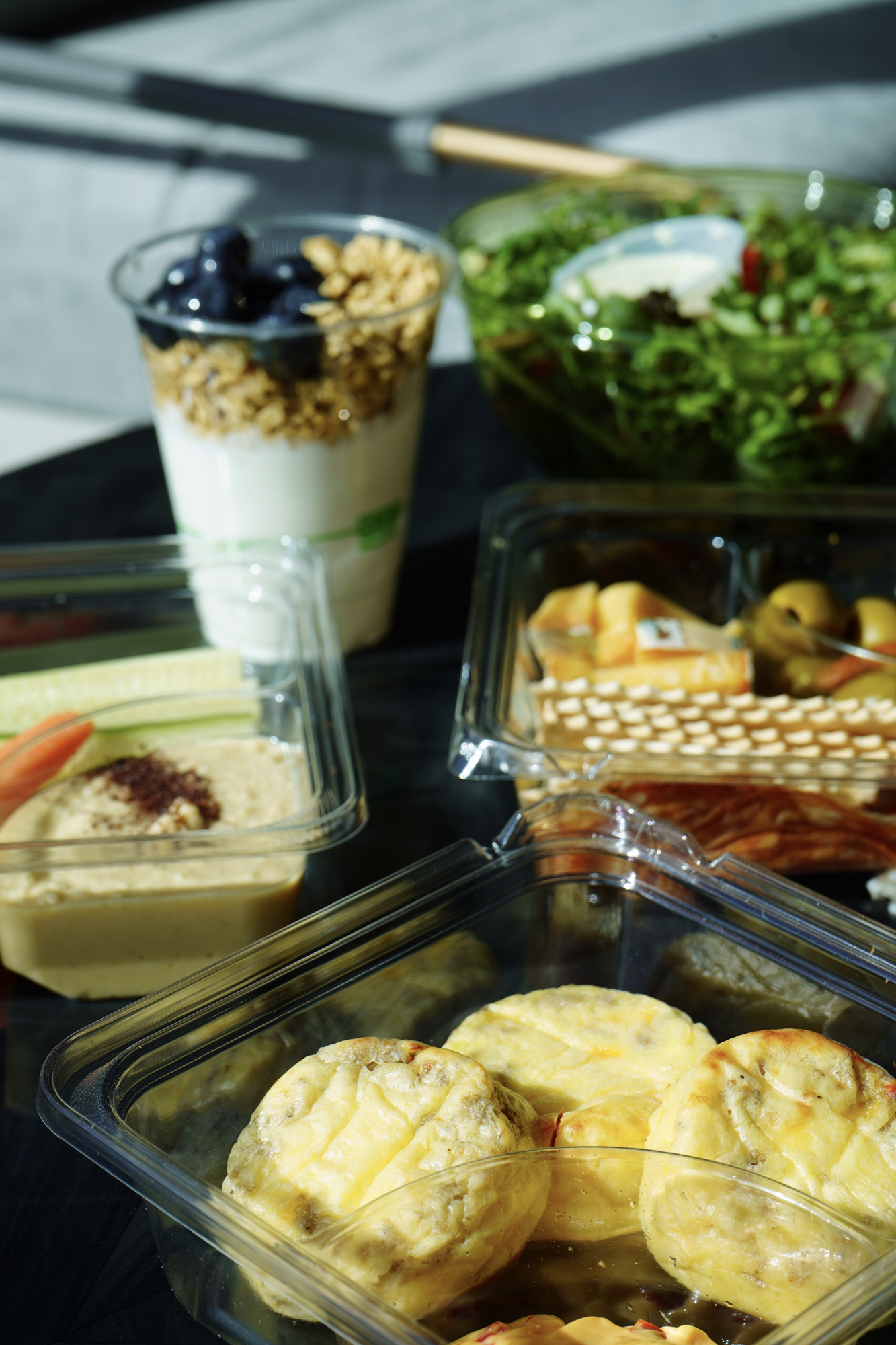 Assorted healthy foods including mini egg muffins in a plastic container, a cup of yogurt with granola and blueberries, a container of hummus with vegetable sticks, a green salad, and a snack tray with crackers, cheese, and peppers.