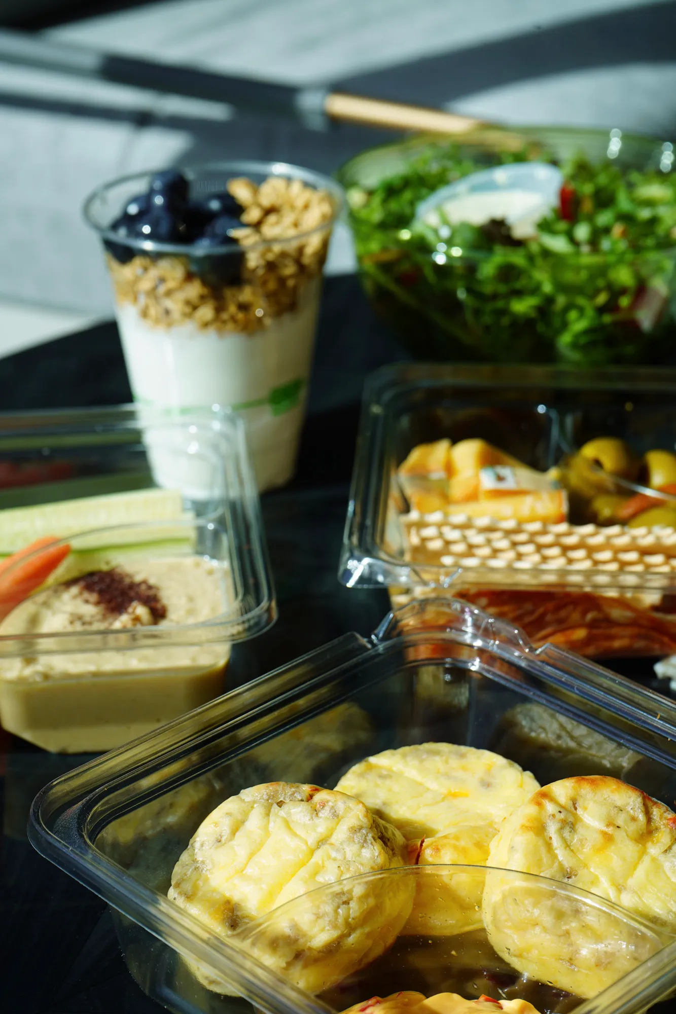 Assorted healthy foods including mini egg muffins in a plastic container, a cup of yogurt with granola and blueberries, a container of hummus with vegetable sticks, a green salad, and a snack tray with crackers, cheese, and peppers.