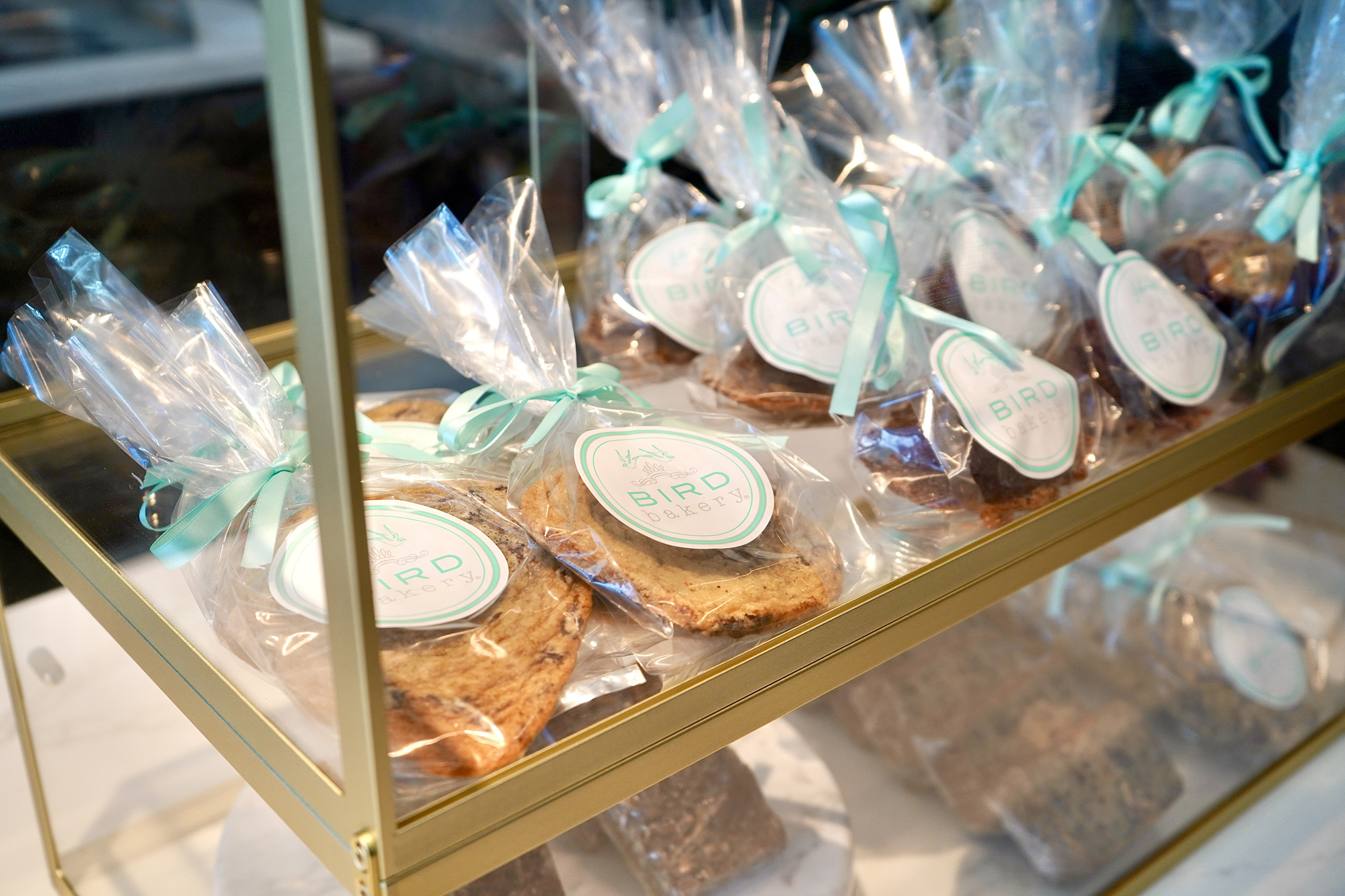 Packaged cookies and baked goods wrapped in clear plastic bags with turquoise ribbons on a gold metal display shelf.