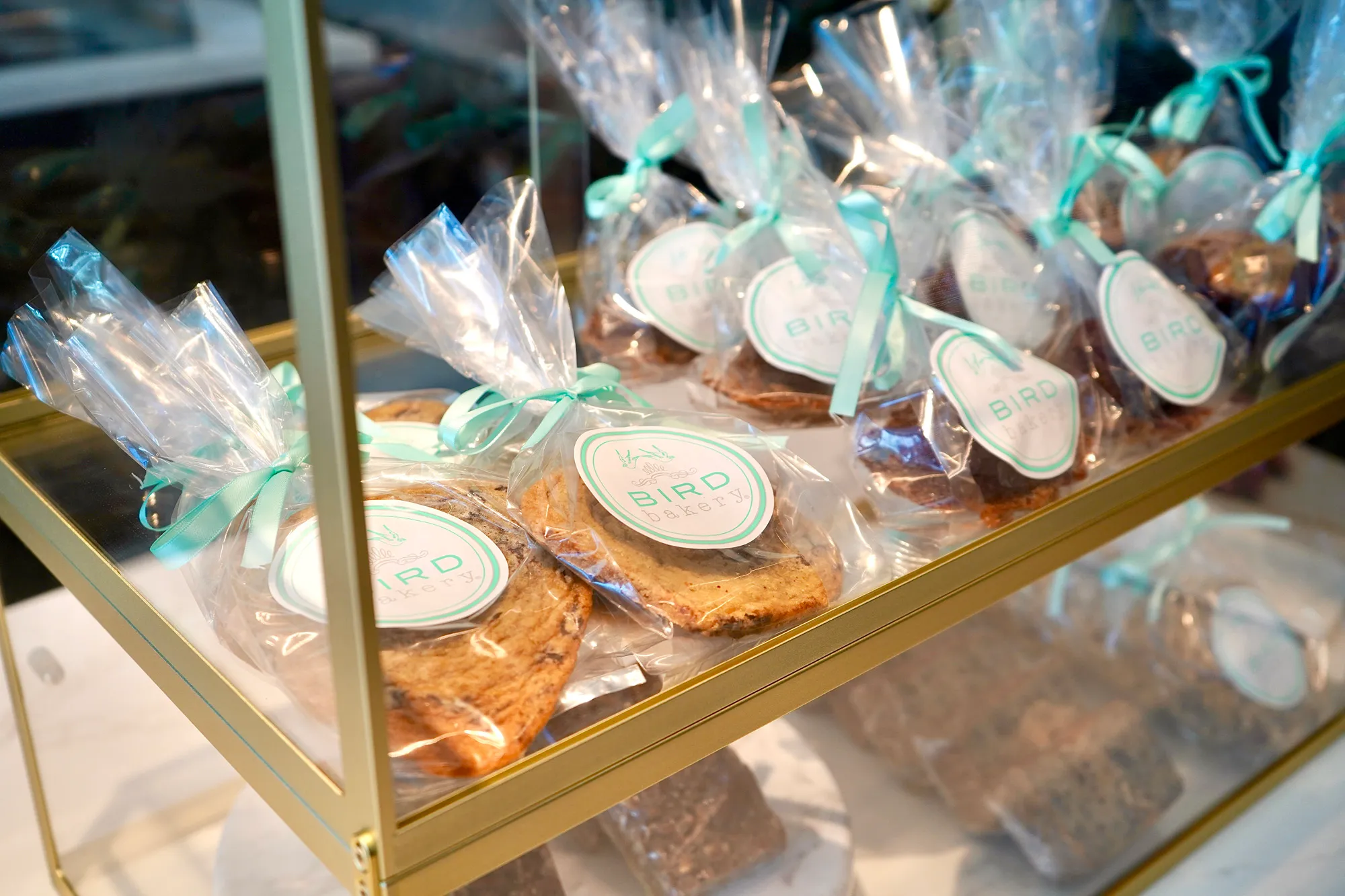 Packaged cookies and baked goods wrapped in clear plastic bags with turquoise ribbons on a gold metal display shelf.