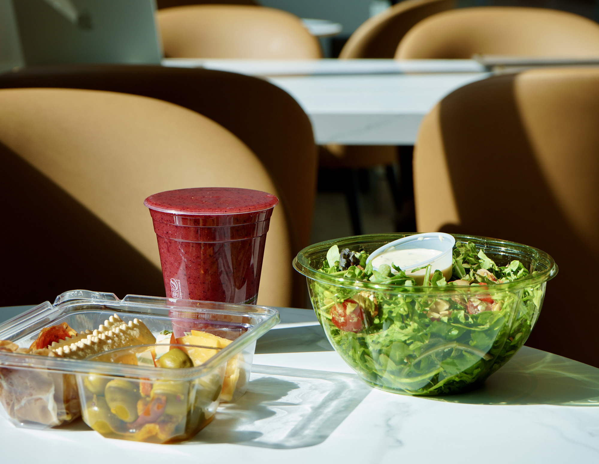 Table with a plastic container of salad with dressing, a smoothie in a sealed cup, and a clear container with olives, cheese, and meats.