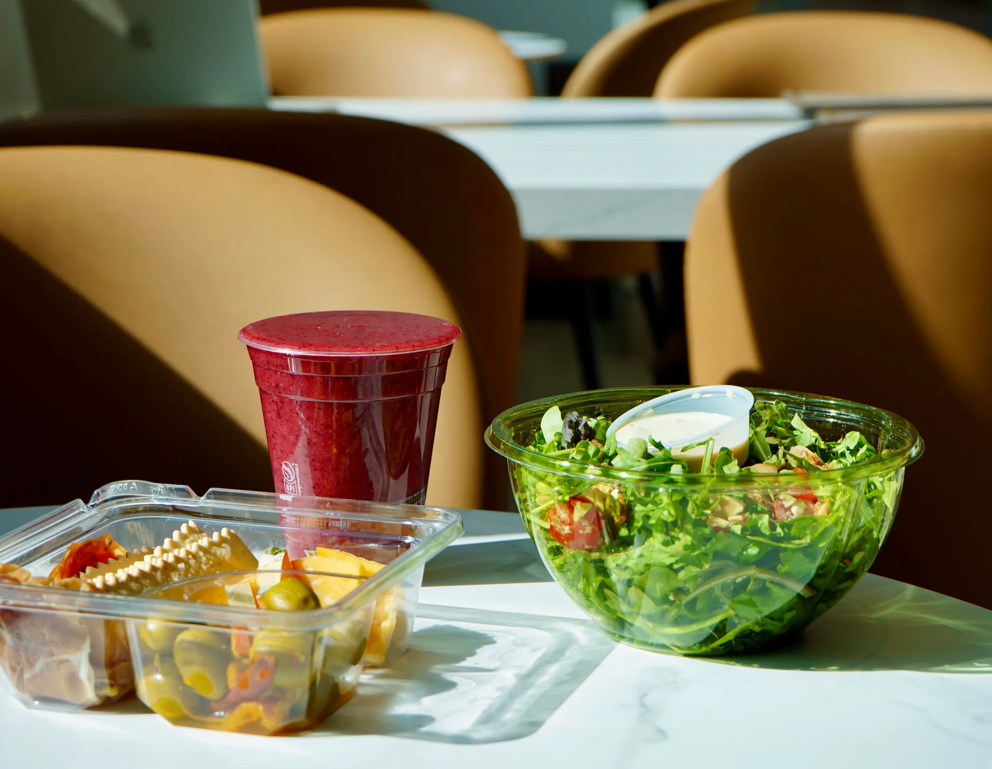 Table with a plastic container of salad with dressing, a smoothie in a sealed cup, and a clear container with olives, cheese, and meats.