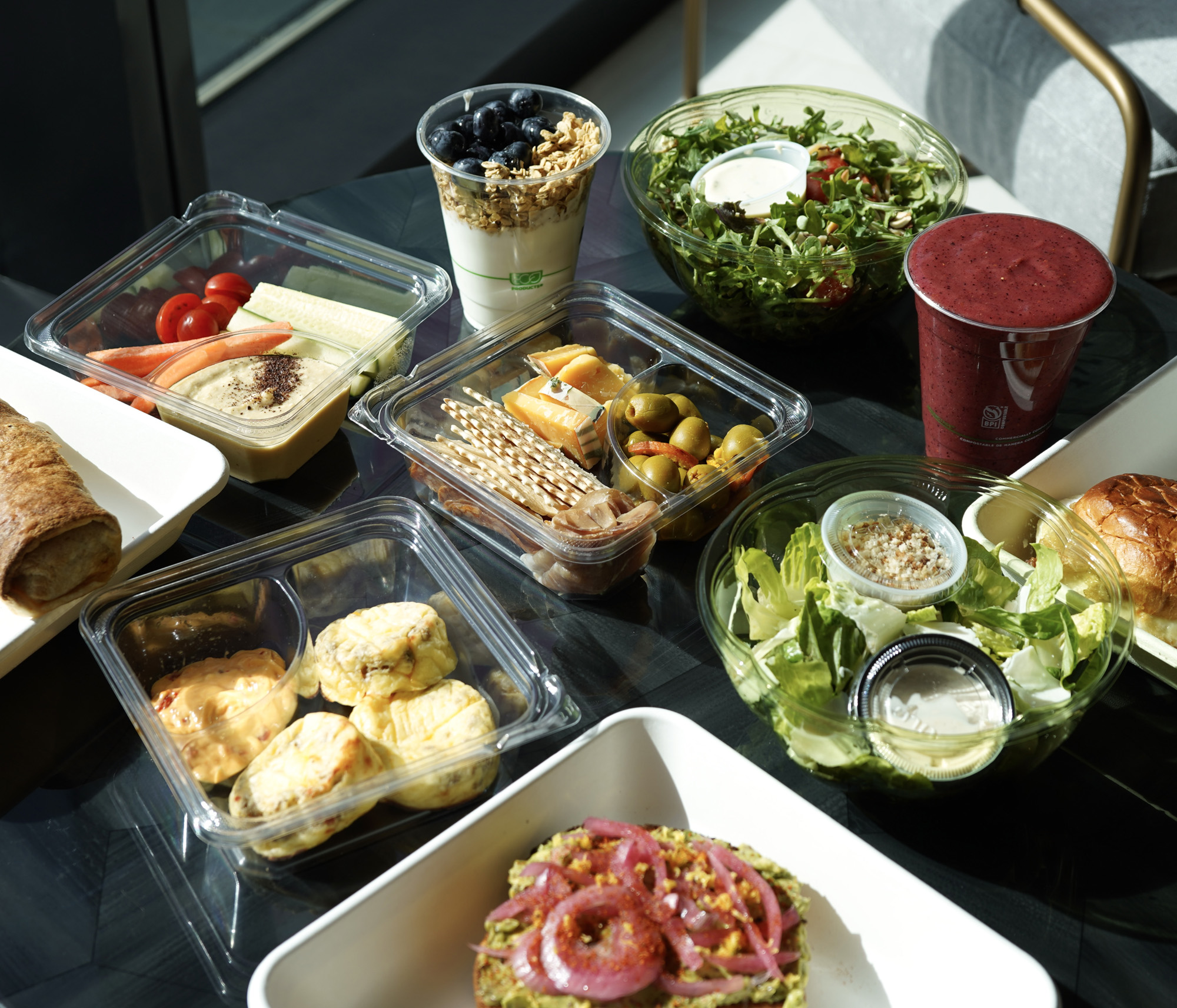 A variety of healthy foods on a table including a mixed green salad, a red smoothie, yogurt with granola and blueberries, a cheese and olive snack pack, a container of vegetables with dip, small quiches with sauce, avocado toast topped with pickled onions, and a sandwich roll.