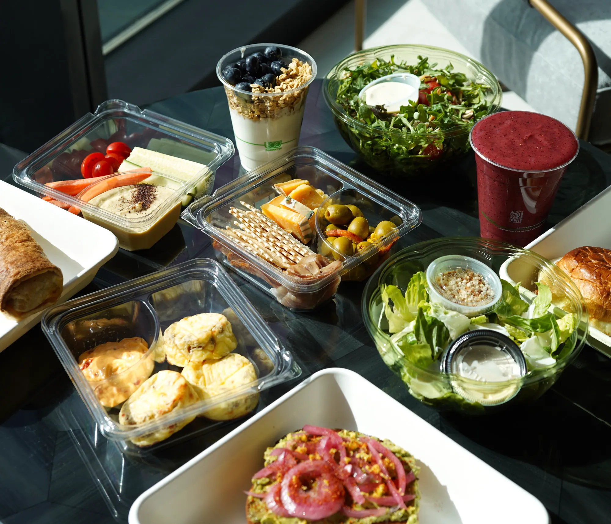 A variety of healthy foods on a table including a mixed green salad, a red smoothie, yogurt with granola and blueberries, a cheese and olive snack pack, a container of vegetables with dip, small quiches with sauce, avocado toast topped with pickled onions, and a sandwich roll.