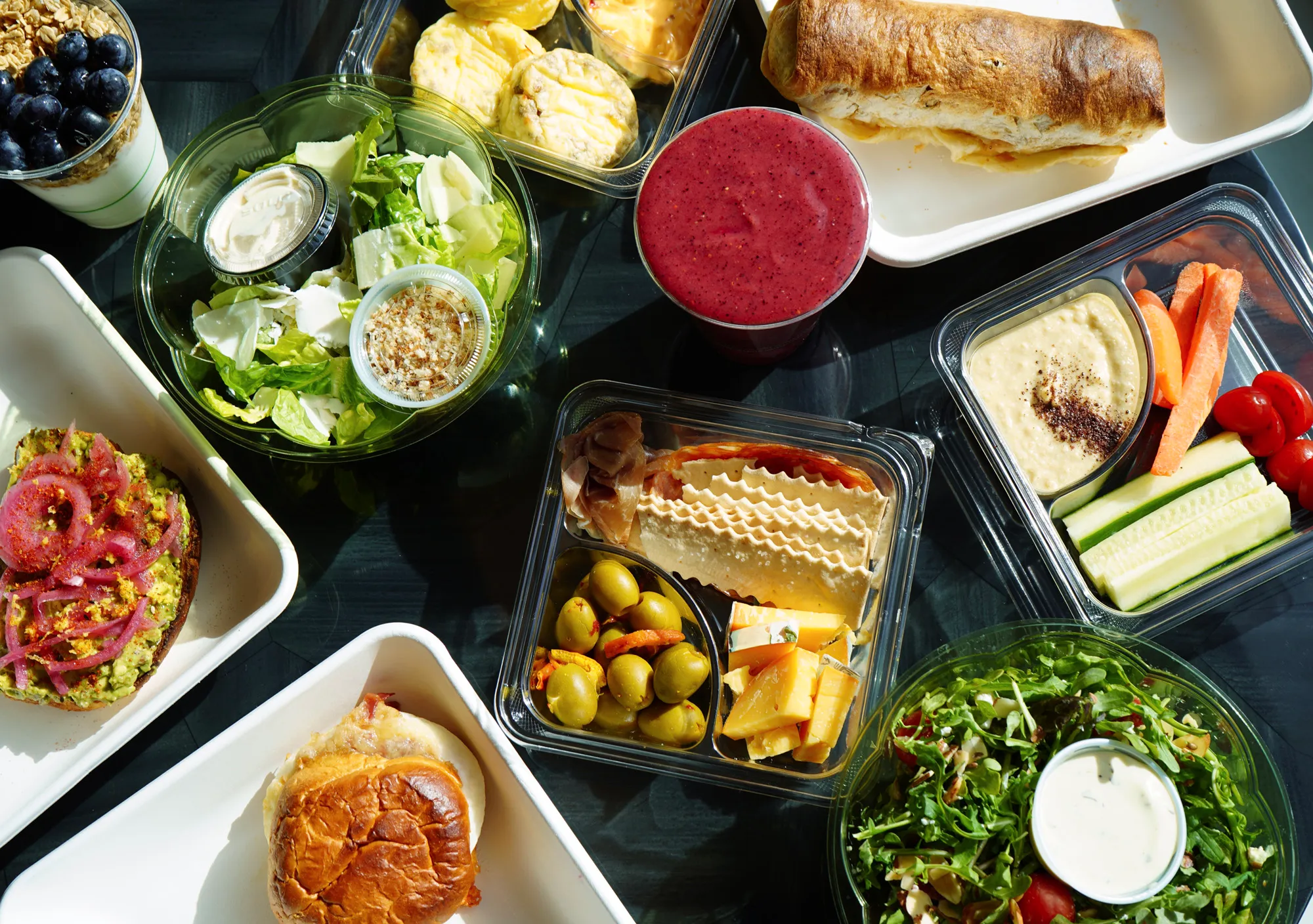 An assortment of healthy meals including a salad with dressing, a sandwich, avocado toast with pickled onions, cheese and olives with crackers, carrot and cucumber sticks with hummus, a berry smoothie, and a cup of granola with blueberries.