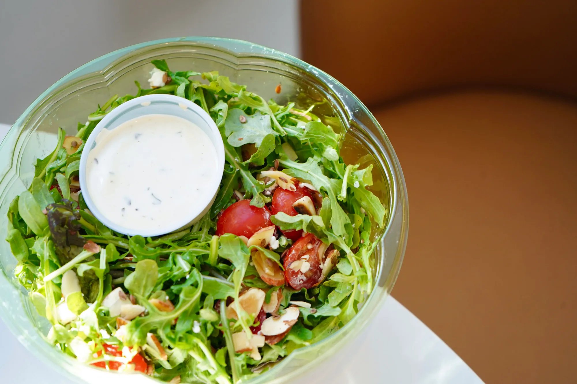 Fresh green salad with cherry tomatoes, sliced almonds, and a cup of creamy dressing in a clear plastic bowl.