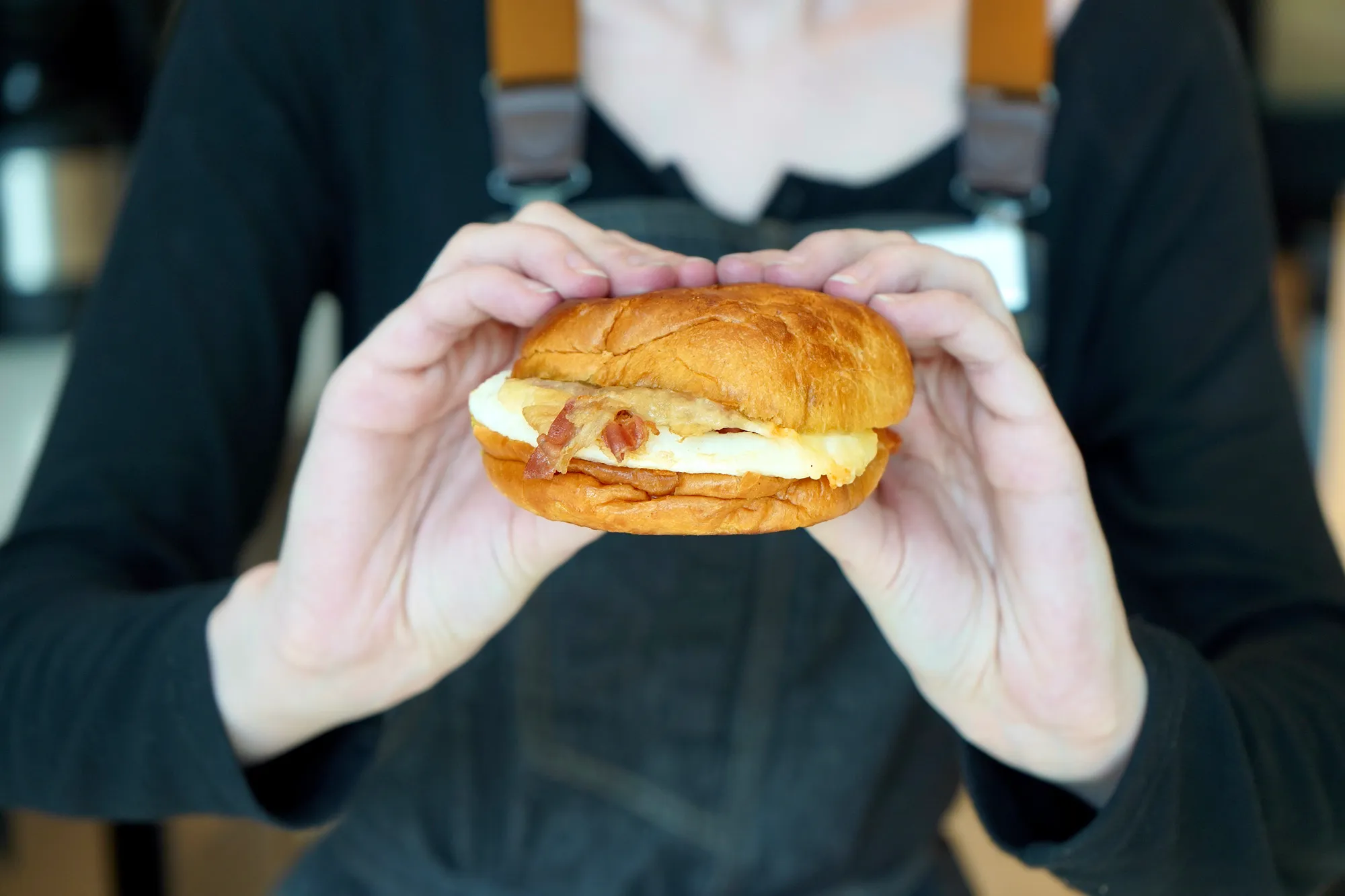 Person holds a breakfast sandwich with egg and bacon in a brioche bun.