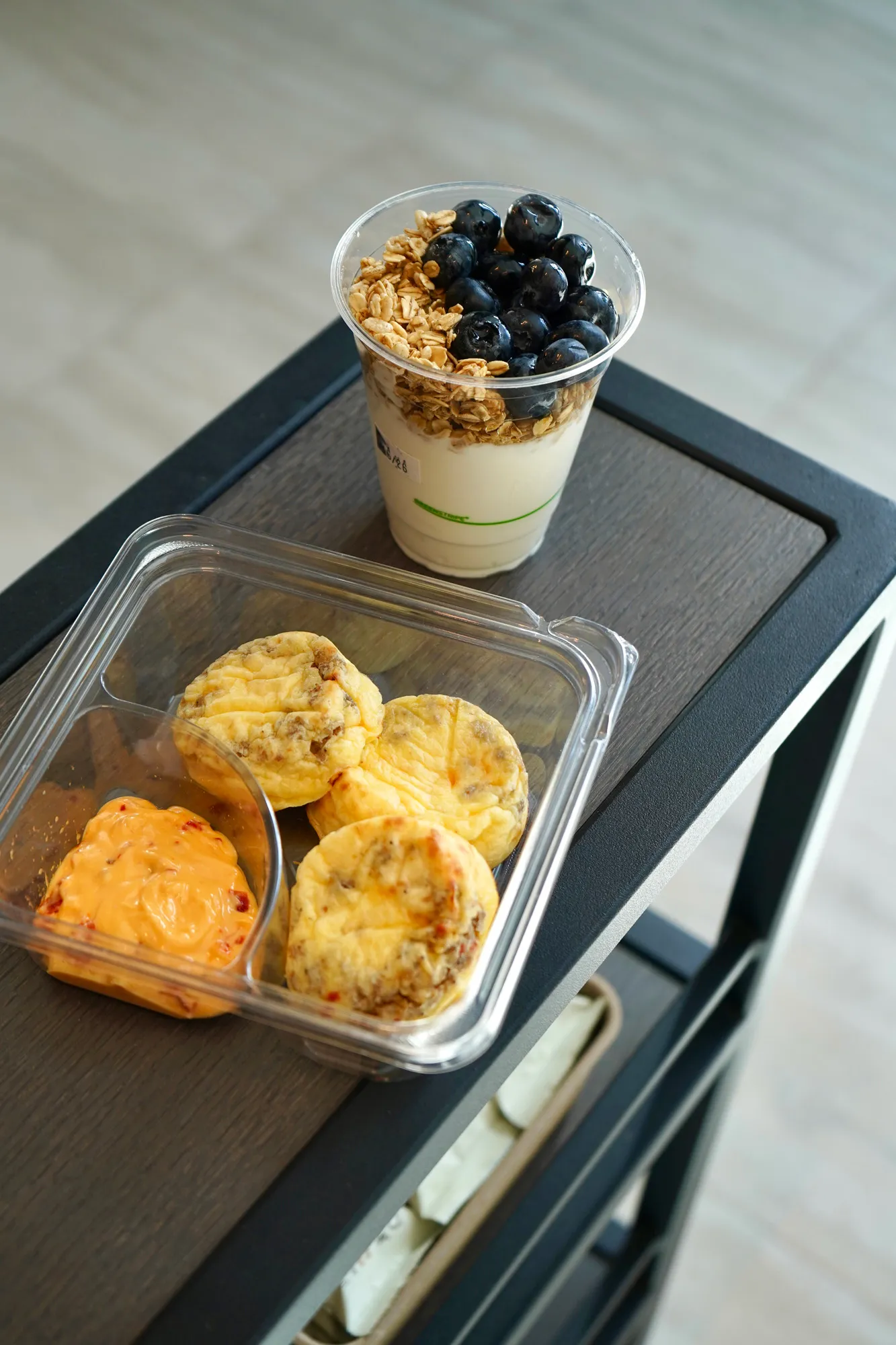 Plastic cup with yogurt topped with granola and blueberries next to a plastic container with three mini egg muffins and orange dipping sauce on a wooden cart.