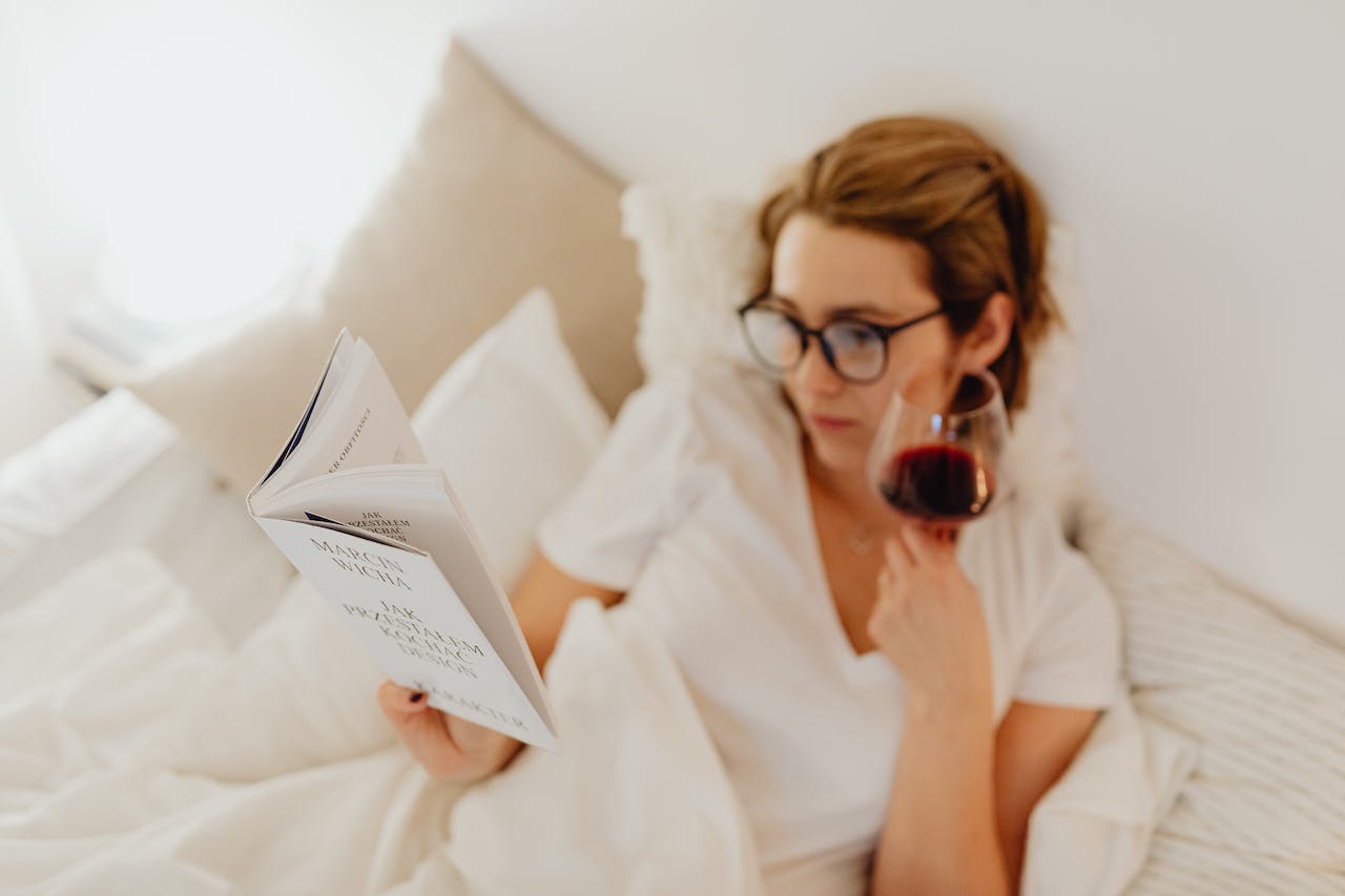 A woman reading a book in bed with a glass of wine
