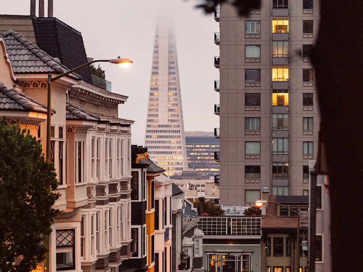 Taken atop one of San Francisco's famous hilly roads, the image captures the city skyline within the fog. A pedestrian walks down the street, which is lined with Victorian-style homes with lit up windows.