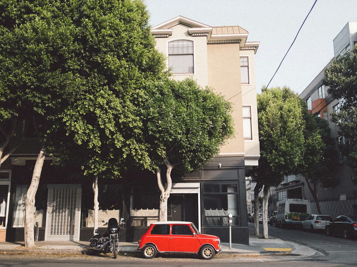In the Hayes Valley neighborhood in San Francisco, a small vintage red car is parked underneath green trees. A classical San Francisco apartment building sits on the corner.