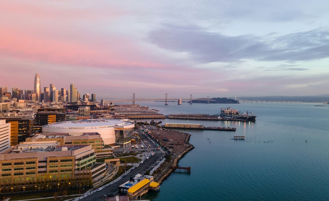 An aerial image of the Mission Bay neighborhood and the Chase Center in San Francisco, with the downtown city center skyline in the background. Taken at sunset, the sky is pink, and in the far distance the Bay Bridge is seen connecting Yerba Buena Island to San Francisco. Piers jut out from the Mission Bay area, connecting to large cargo ships in the waters of the bay.