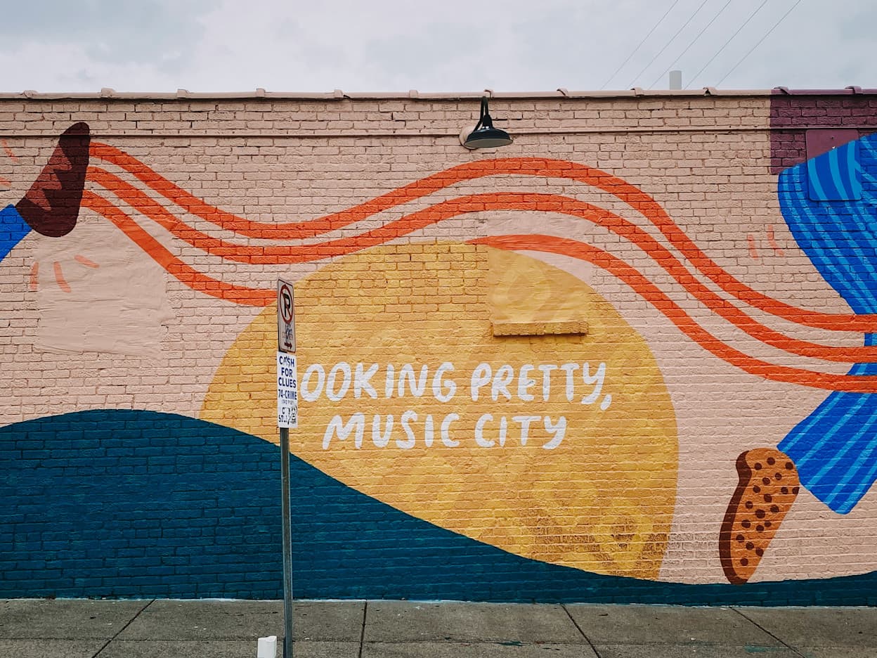 An image of a mural that says "Looking Pretty, Music City" painted on a brick wall in Nashville. Painted in orange, yellow, and blue colors, the words in white pop out in the mural and refer to Nashville's musical reputation.
