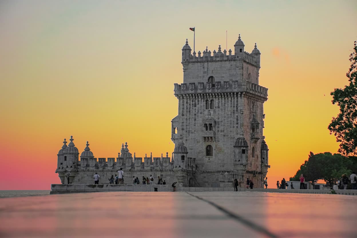 An image of the famous and historical castle in Belém, Lisbon in Portugal. There is a bright sunset in the background ranging from bright yellow to deep oranges, making the castle's silhouette pop out against the sky.