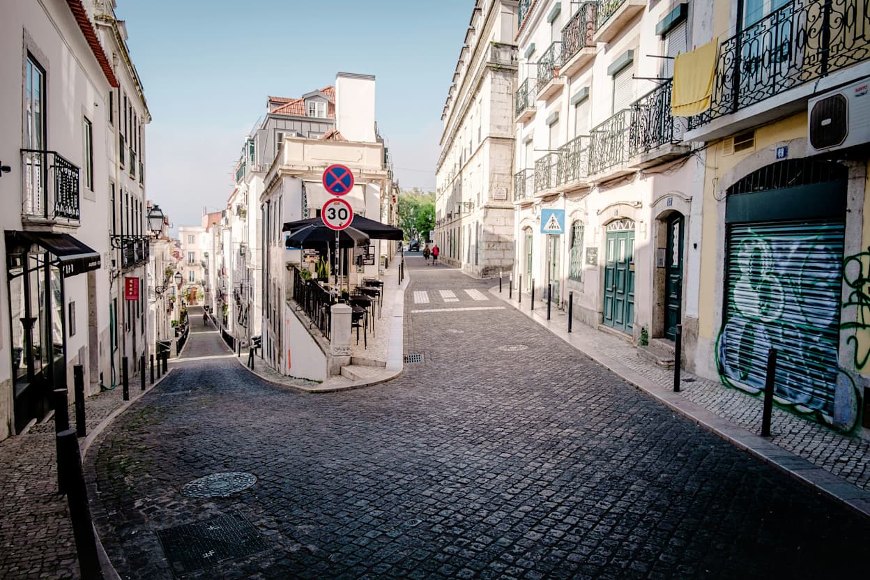A picture of a winding cobblestone road that completes a full U-turn in the Bairro Alto neighborhood in Lisbon. White buildings line the streets. and there is street art on the garage doors. Iron balconies are also along the buildings.