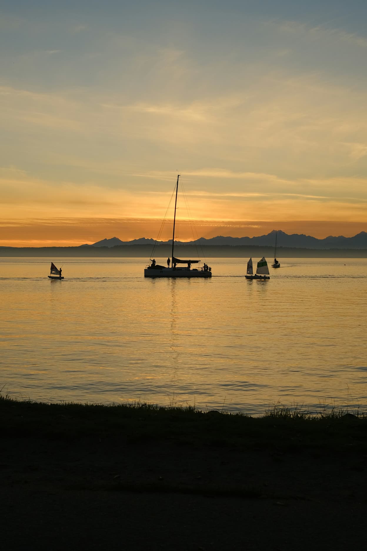 An image taken at sunset capturing the silhouette of a sailboat in the water in Ballard, Seattle. The orange sunset reflects on the water, and the mountains are visible in the horizon.
