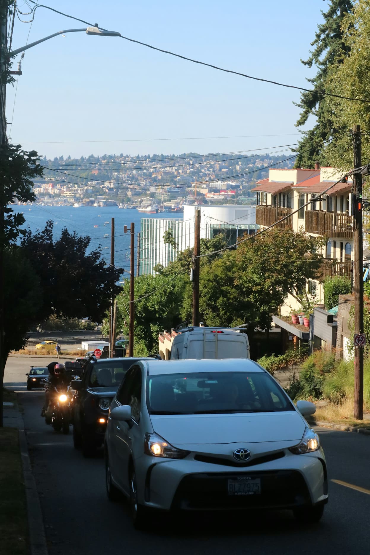 Taken in the street in Capitol Hill, Seattle. Cars drive up the hill, and trees and houses line the road. Beyond the road you can see the bay of water, and another hill of land on the other side of the water. Lush green trees fill the land on either side.