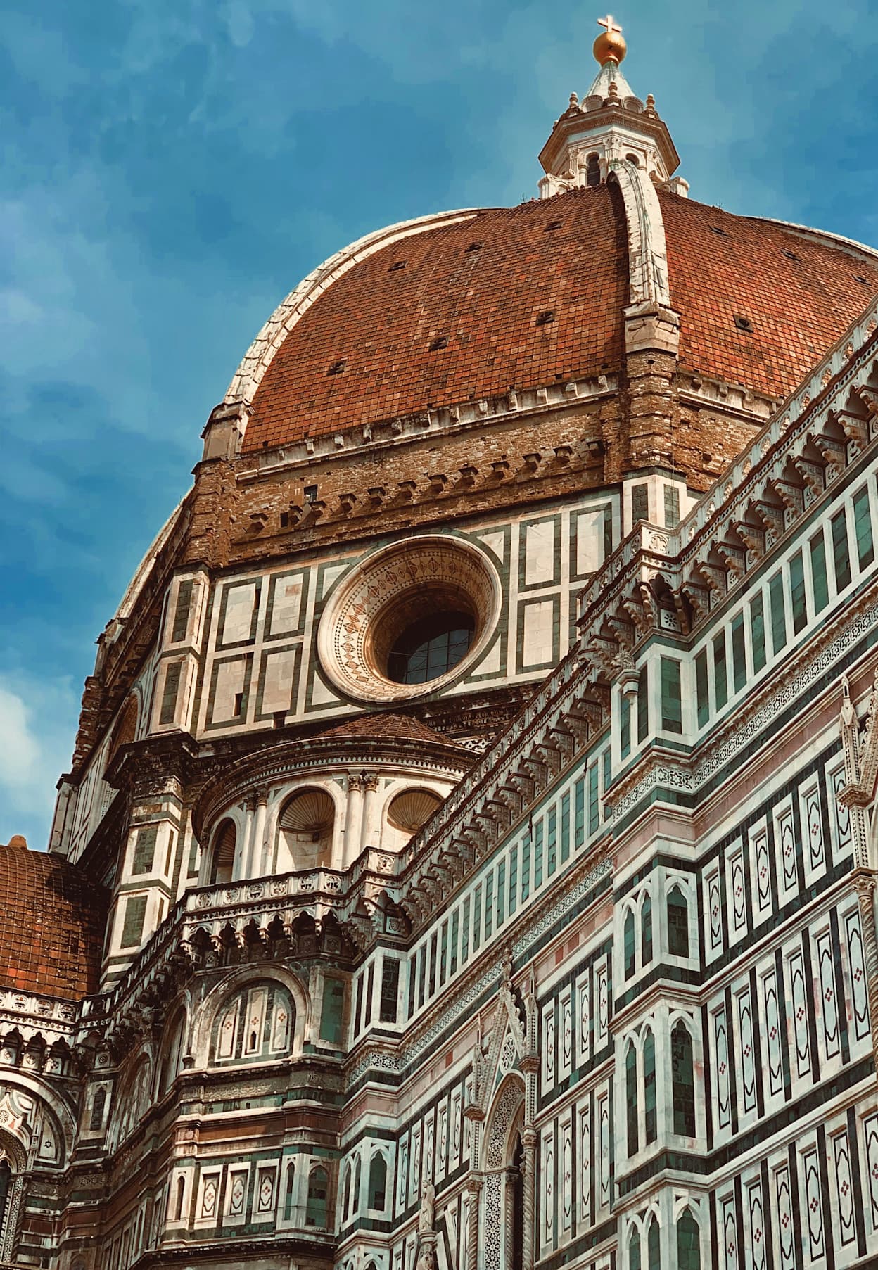 A close-up image taken from the street of the famous Duomo Cathedral in Florence, Italy. The dark and light tile exterior creates a geometric pattern along the building and the terracotta dome on top pops against the blue sky. 
