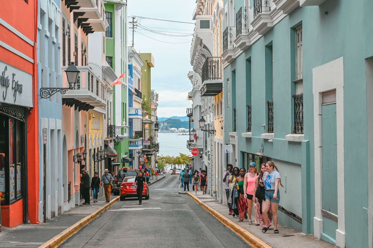 An image was taken in the middle of a road in Old San Juan, Puerto Rico, of bright-colored houses along a narrow road. People walk along the thin sidewalks, and at the end of the road, you can see a body of water.
