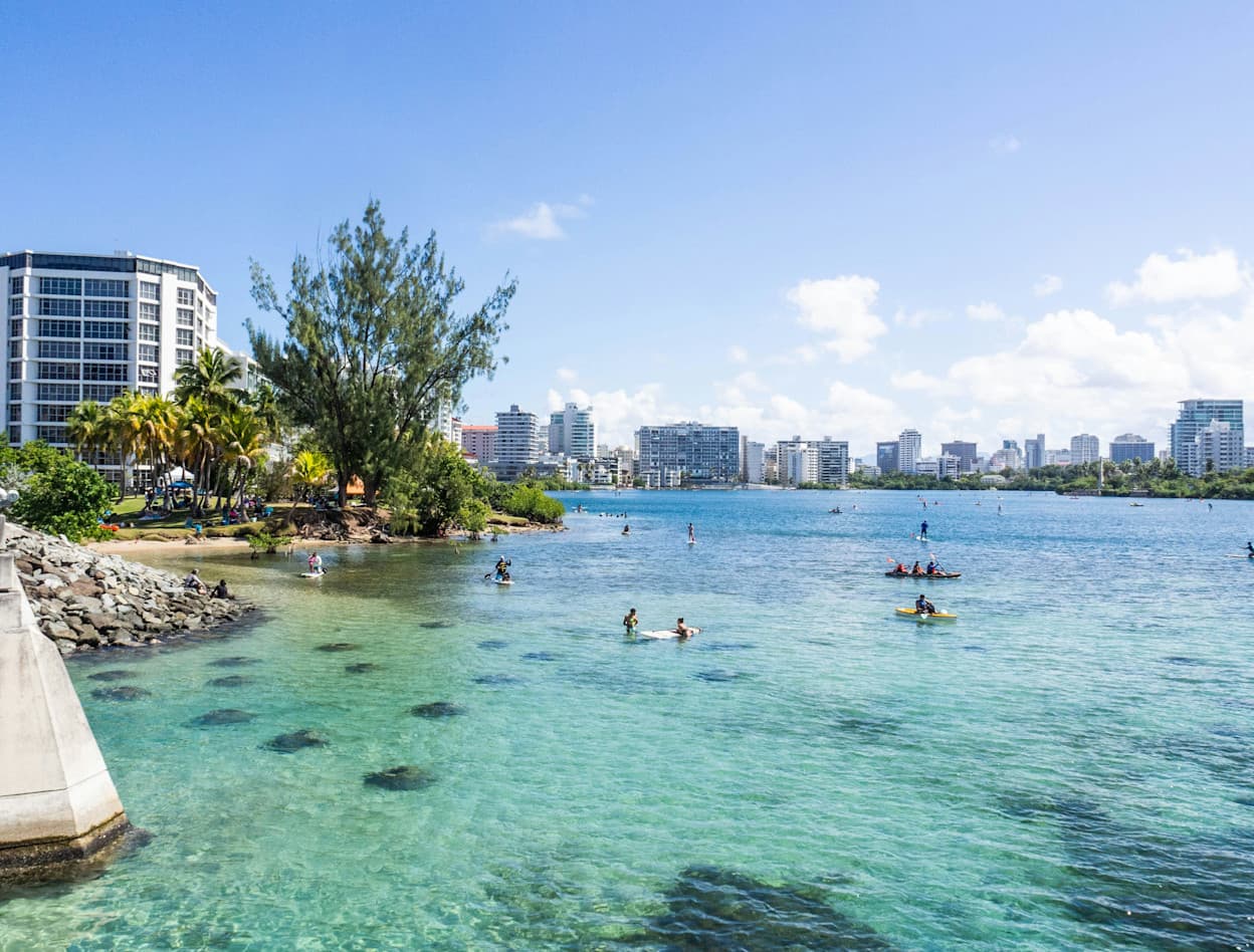 People swim in the clear waters at Condado Beach in San Juan, Puerto Rico. Tall buildings line the beach. Some rocks are seen on the bottom of the ocean floor where people swim.