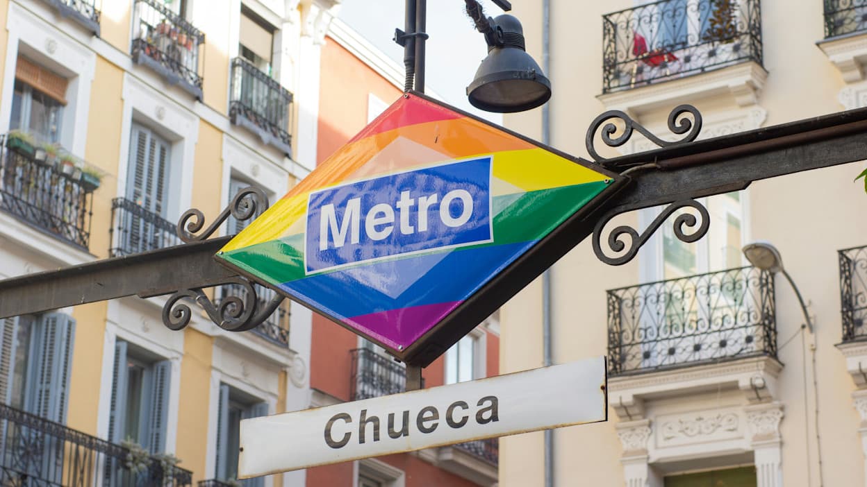 An image of the Chueca metro stop, which is in a rainbow pattern to symbolize the LGBT history the Chueca neighborhood holds. Behind the sign, you can see apartment buildings.