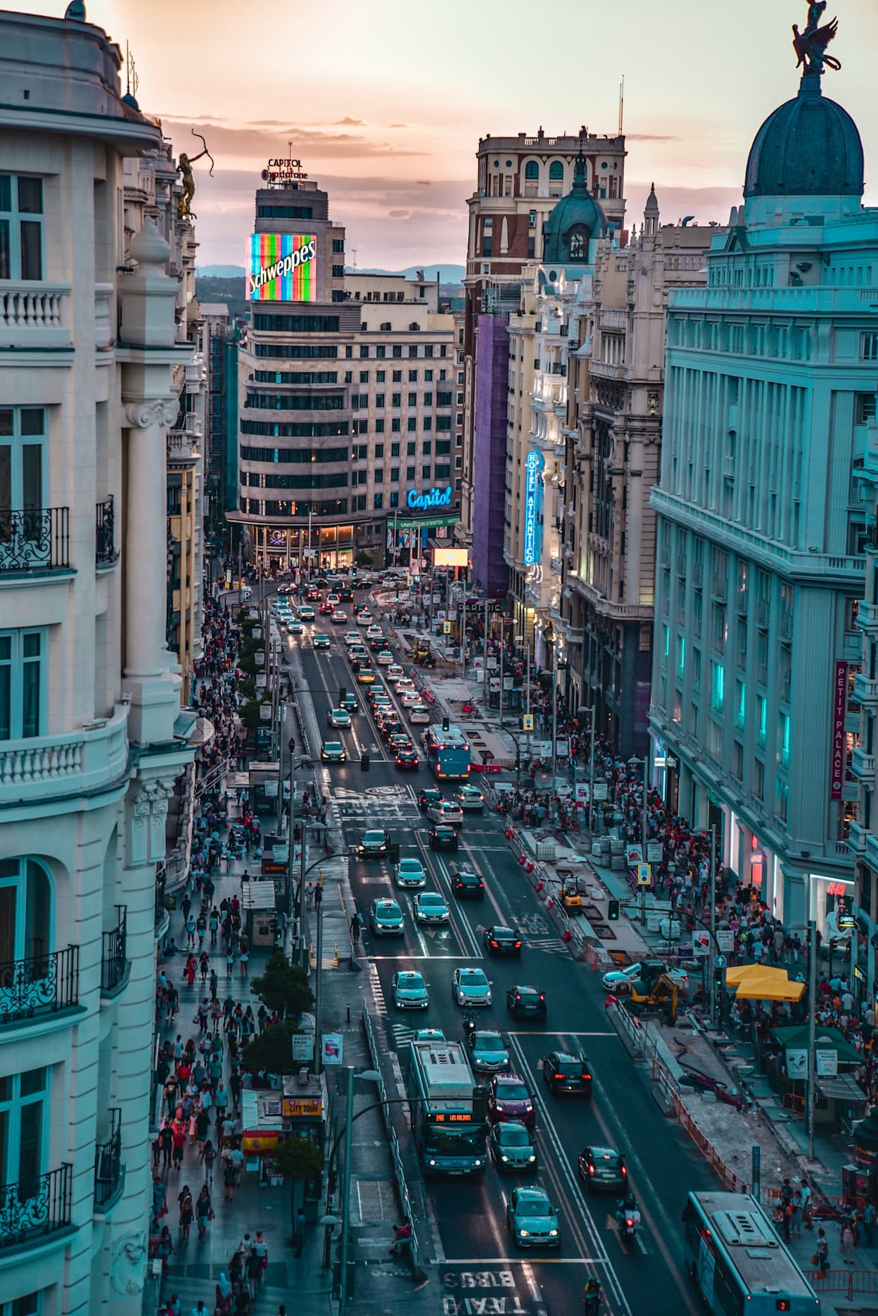 An image of the bustling Gran Vía street in the Sol Gran Vía neighborhood in Madrid, Spain. Cars fill the street, and there are many people walking. Many tall buildings line the road.
