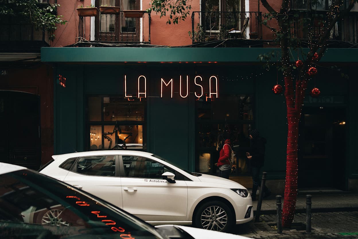 An image of a storefront in Malasaña, Madrid that says "LA MUSA" in light up letters. A white car is parked in front of the building on the street.