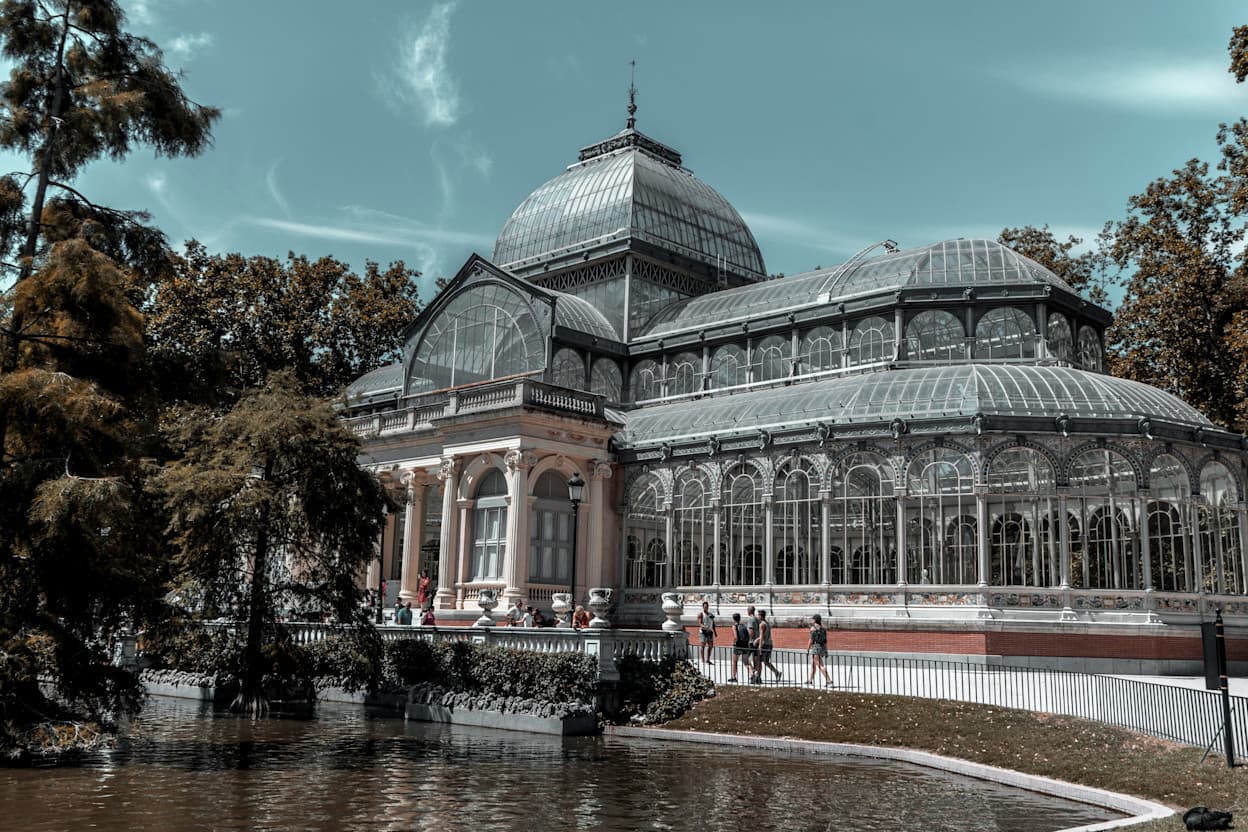 An image of the Palacio Cristal in Retiro Park in Madrid, Spain. The all-glass palace is see-through and is surrounded by trees. It sits on the side of a pond.