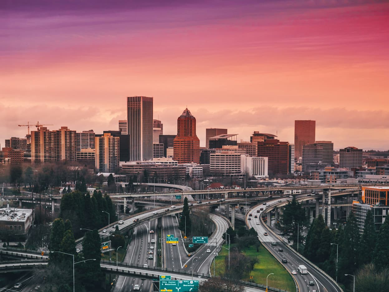 An image taken from above a freeway with multiple freeways in the foreground, and the Downtown Portland city skyline in the background against an orange and purple sunset.