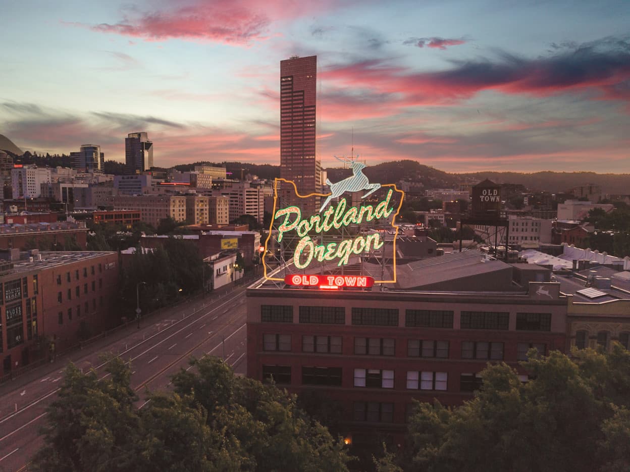 An image of a light-up sign on top of a building in the shape of the state of Oregon that says "Portland, Oregon." In the background, the Portland skyline is seen against a cloudy pink sunset.
